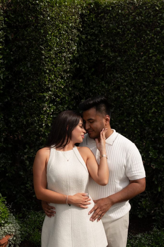 Engaged couple posing in front of lush greenery at the Dallas Arboretum. Woman in a sleeveless white dress caresses her partner’s face as they share an intimate moment, both dressed in light neutral tones