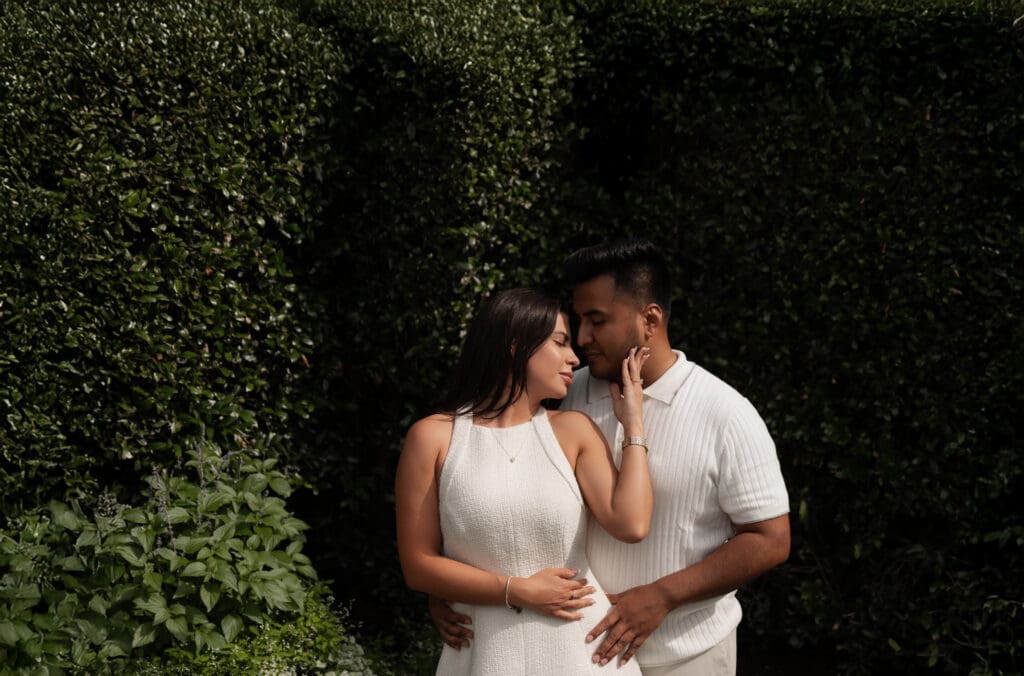 Romantic engagement photo of a couple embracing in front of a green hedge, captured in natural light by a professional photographer in Dallas, Texas.