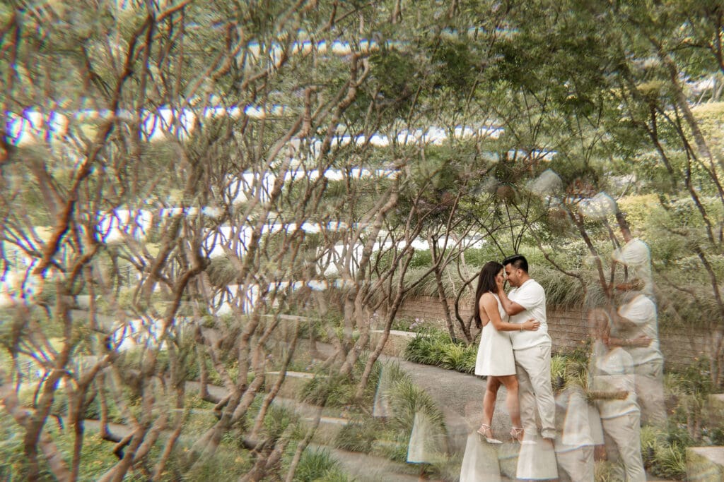 Engagement photo of a couple embracing under trees at the Dallas Arboretum, with creative prism effect adding dreamy reflections. Woman in white dress, man in light outfit, romantic garden setting.
