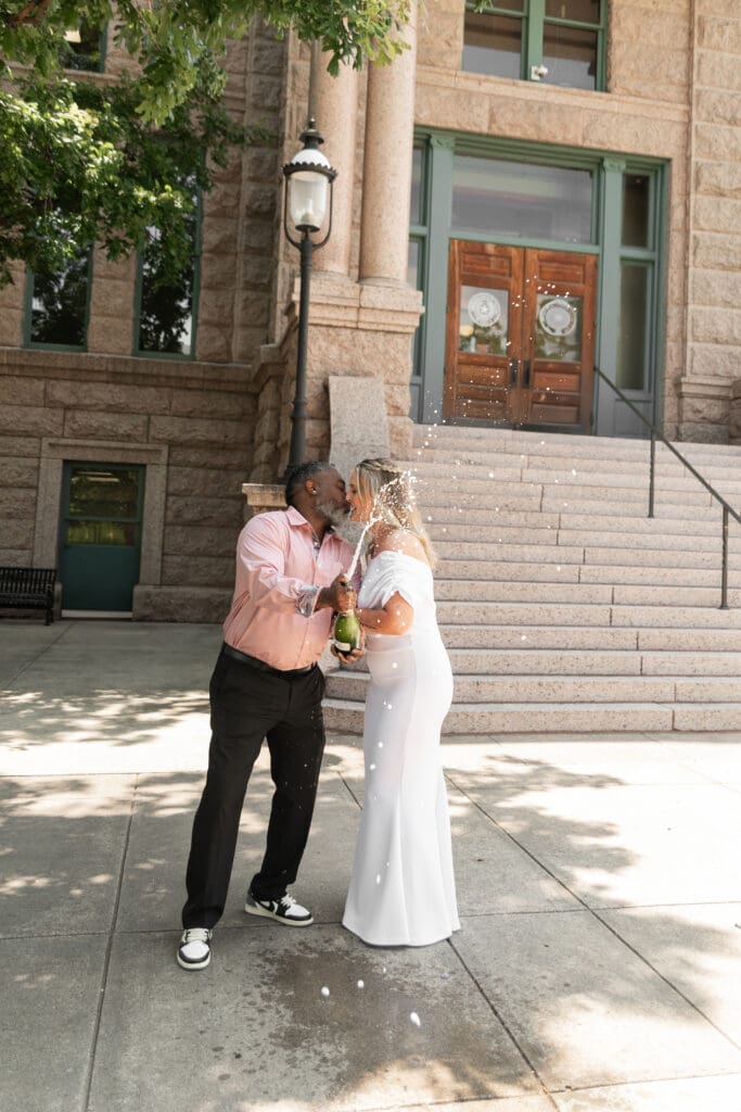 Bride and groom celebrating their intimate Fort Worth courthouse wedding by popping champagne outdoors.