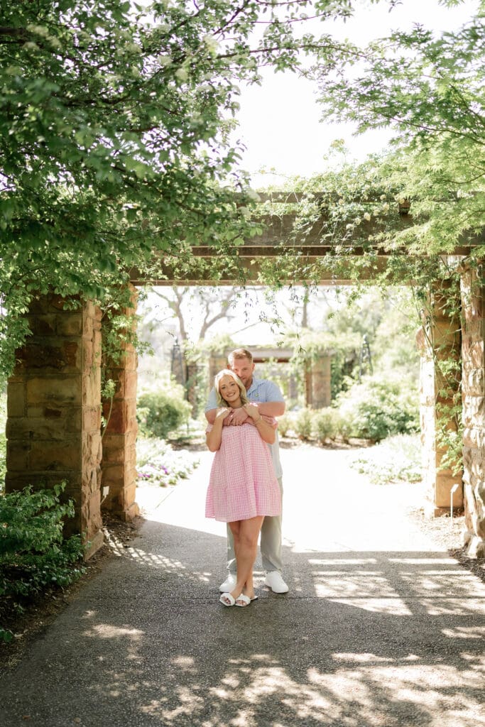 Woman and man cuddling under the archway at the Fort Worth Botanic Gardens during their engagement session