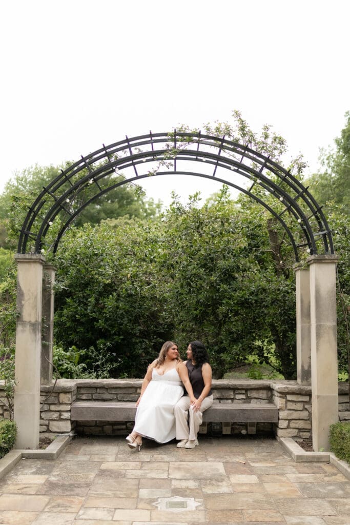 Two brides sitting on a bench and snuggling up at the Fort Worth Botanic Garden during their engagement session