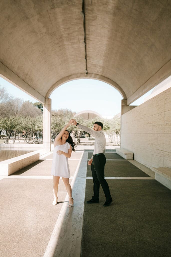 Engaged couple at Kimbell Art Museum, Fort Worth, holding hands and dancing under an arched hallway