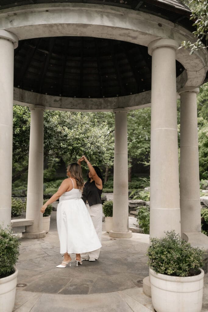 Two women dancing under a gazebo during their Fort Worth engagement session at the Fort Worth Botanic Garden