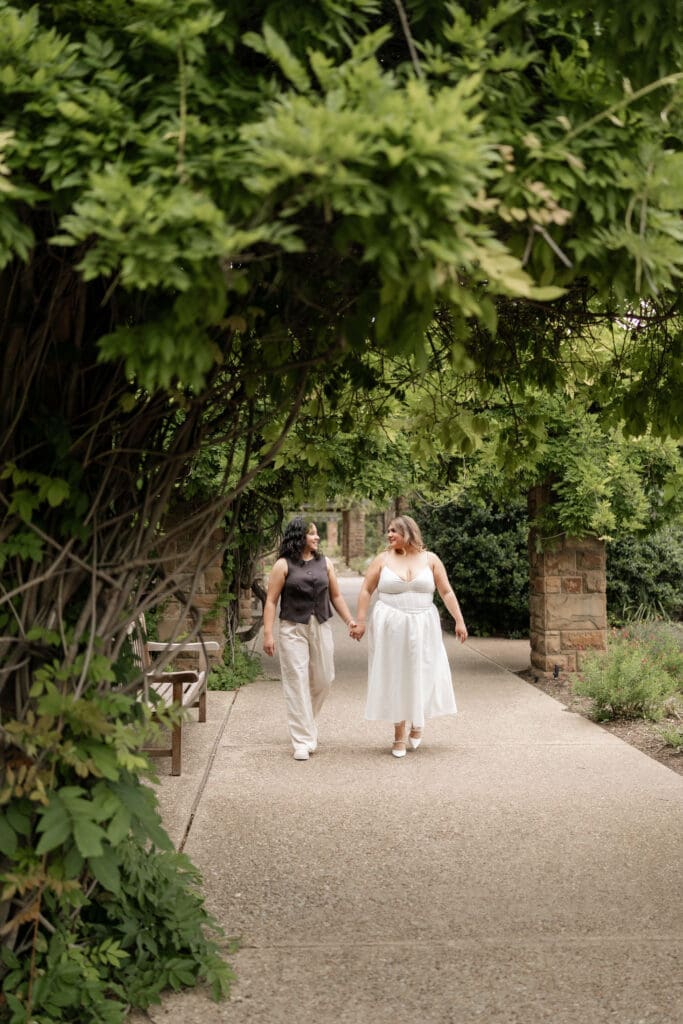Two women walking and holding hands under the wisteria tunnel during their Fort Worth engagement session at the Fort Worth Botanic Garden