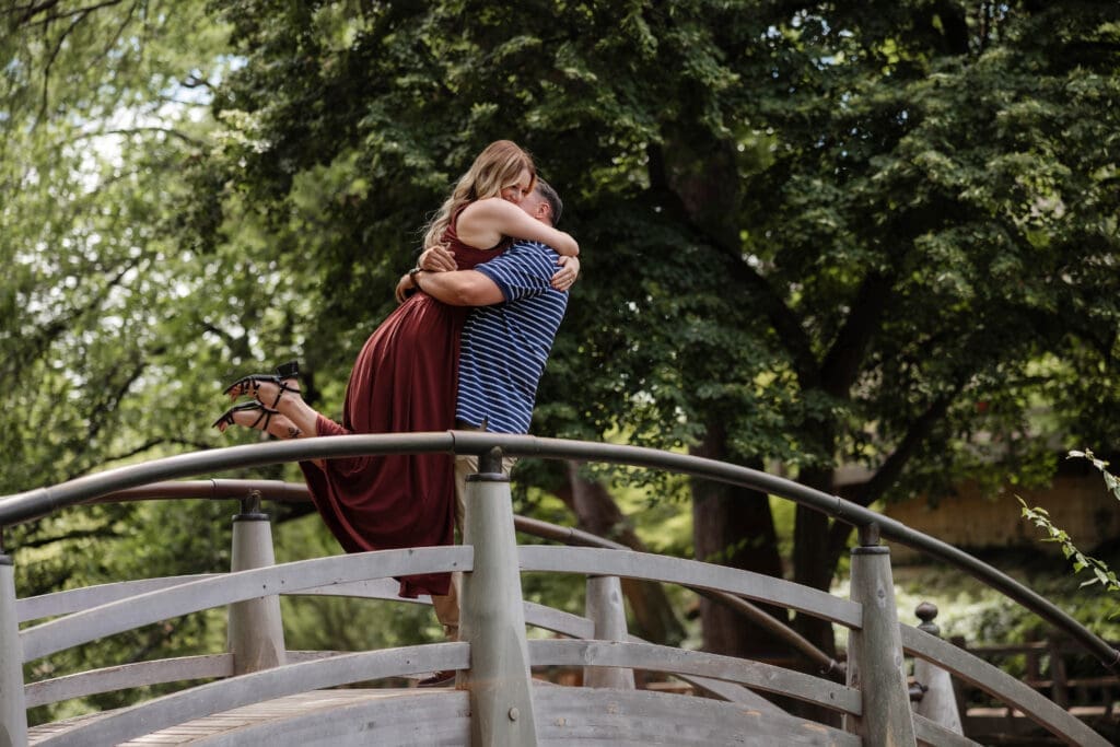 Man and woman embracing in a big hug on the bridge in the Japanese Garden at Fort Worth Botanic Garden after she said yes to his proposal