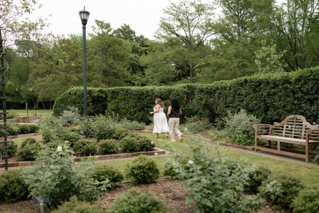 Two women playfully chasing each other in the garden during their Fort Worth engagement session at the Fort Worth Botanic Garden
