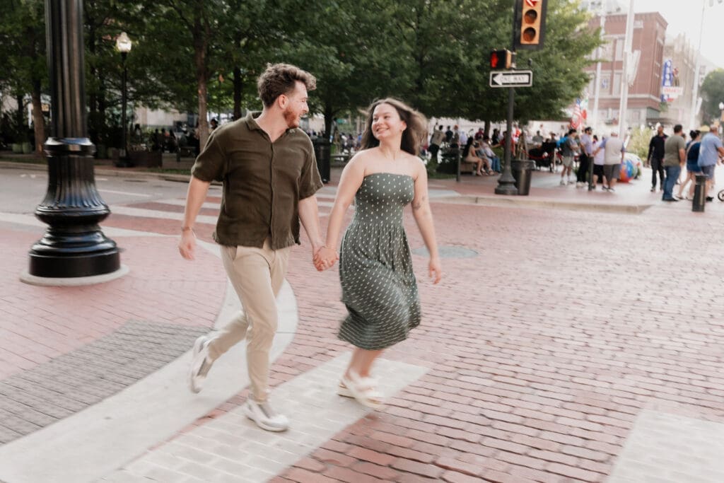 Man and woman running across a crosswalk in Sundance Square, Fort Worth, TX during their engagement photo session