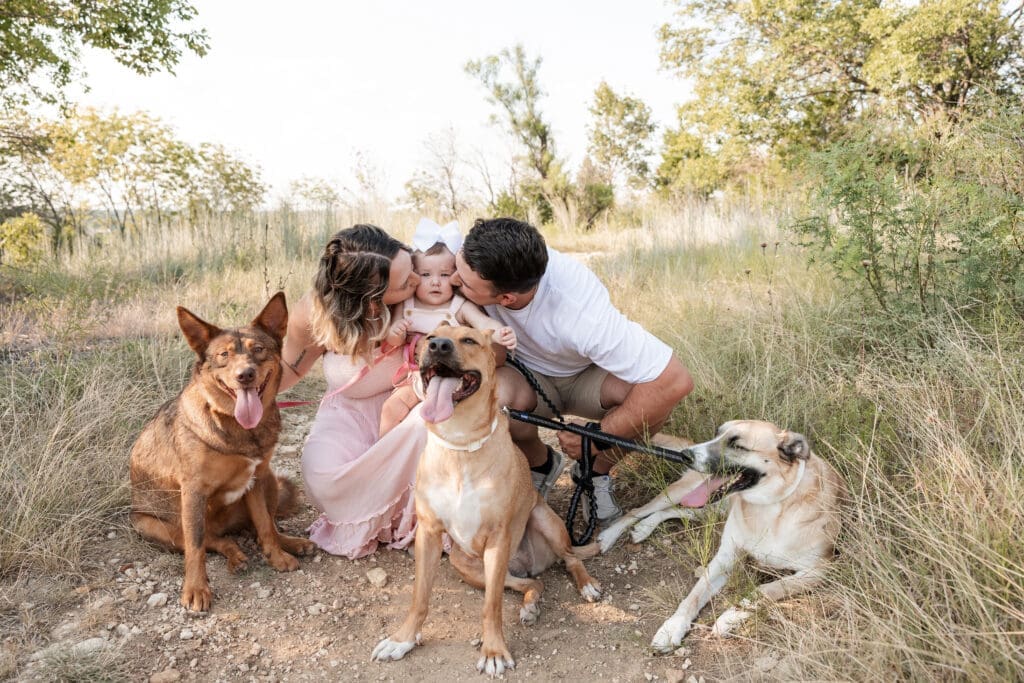 Husband and wife kissing their baby while sitting in the field at Tandy Hills in Fort Worth with their dogs for their family photography session