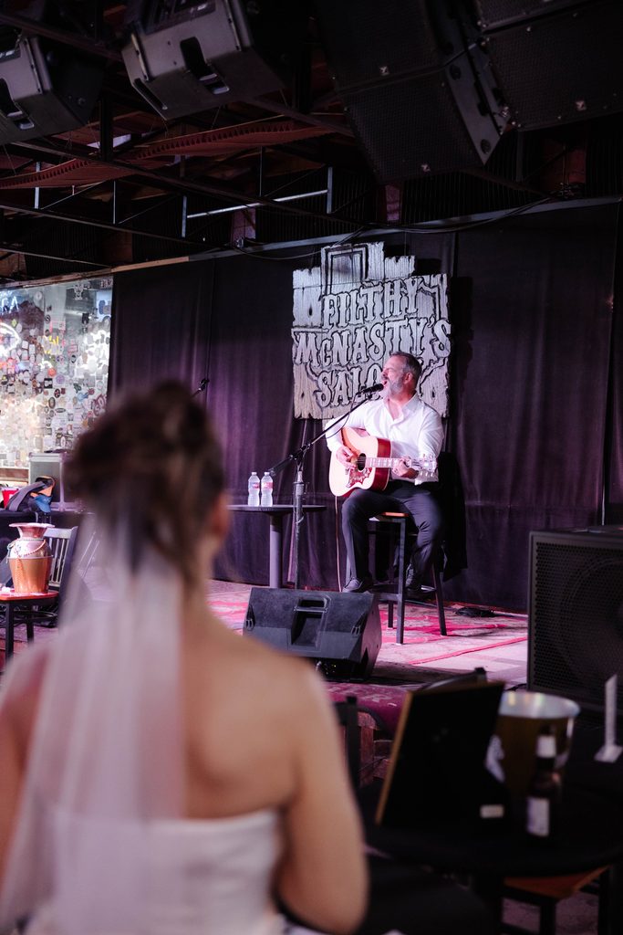 Jason singing and playing guitar on stage at Filthy McNasty's Bar in the Fort Worth Stockyards while his bride watches from a distance