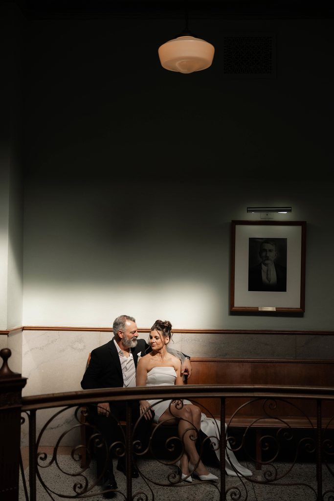 Bride + Groom sitting on wooden bench at the Tarrant County Courthouse posing for a moody portrait during their adventure elopement in Fort Worth